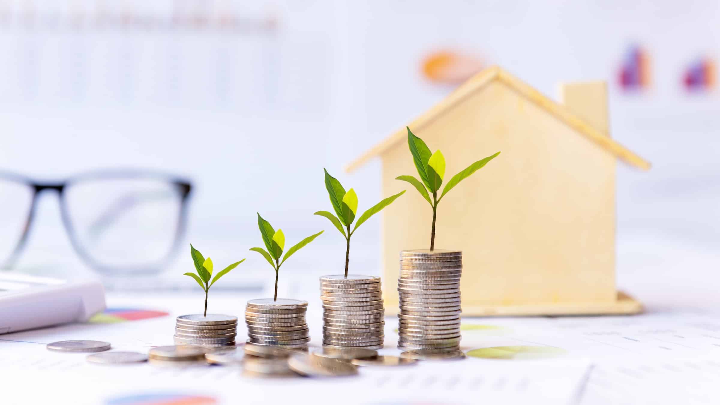 Investment concept showing sequentially larger stacks of coins with plant seedlings growing from them in front of a model house