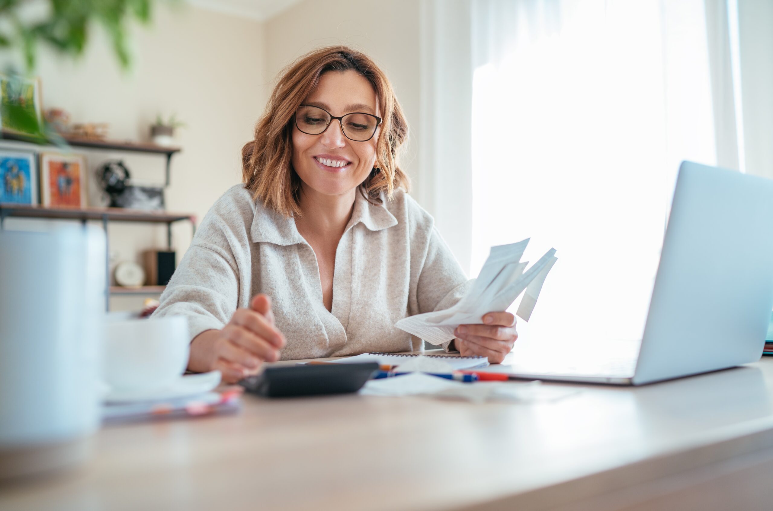 Beautiful,Middle-aged,Woman,In,Glasses,And,Paper,Bills,Joyfully,Laughing woman looking at her bills and laptop